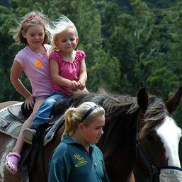 Duas meninas montando um cavalo e uma menina caminhando ao lado dele